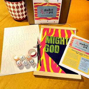 May include: A paint-by-number kit featuring a cardboard box with a Strokes of Joy label, a pre-printed canvas sheet, a finished canvas with wooden hangers displaying the text MIGHTY GOD, six numbered paint pots, two brushes, and an instruction card.