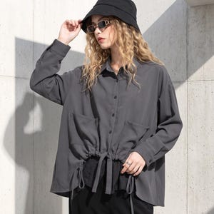 Puede incluir: Una camisa de botones gris carbón de manga larga con dos bolsillos delanteros y una cintura con cordón ajustable y lazos colgantes. La camisa se combina con un sombrero de pescador negro, gafas rectangulares y pantalones negros.