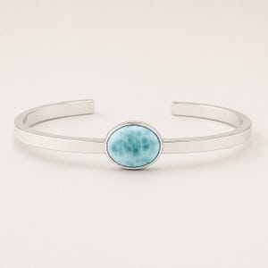 May include: A silver cuff bracelet featuring a smooth, polished metal band with an open design. Centered on the front is a single, oval-shaped Larimar gemstone displaying a marbled pattern of light blue and white tones with a polished finish.