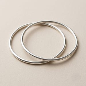Peut inclure: Deux bracelets joncs circulaires en métal argenté, lisses et polis, dotés d'un profil fin et arrondi. Les bracelets sont superposés, mettant en valeur une surface métallique réfléchissante et un design simple en anneau continu sans ornement.