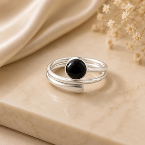 May include: A silver bypass ring featuring a polished, round black stone set in a smooth bezel. The band is crafted from a continuous piece of silver that wraps around the finger, creating an open, adjustable design with a sleek, metallic finish.
