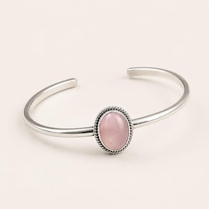 Può includere: Un bracciale rigido in argento con una pietra ovale rosa chiaro al centro. La pietra è incastonata in un bordo metallico decorativo a corda intrecciata, e la fascia del bracciale è in argento liscio e lucido con un design aperto sul retro.