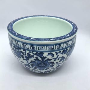 May include: A round ceramic bowl featuring a white base with intricate blue floral patterns and scrolling vines. The rim is decorated with a blue and white geometric Greek key border and a thin band of stylized leaf motifs around the top edge.