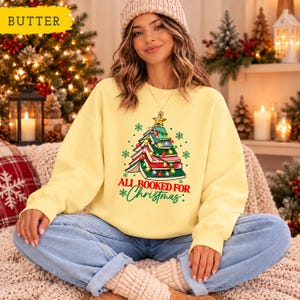May include: A pale yellow long-sleeve sweatshirt featuring a graphic of a Christmas tree made of stacked books, topped with a gold star and decorated with colorful string lights and snowflakes, with the text "ALL BOOKED FOR CHRISTMAS".