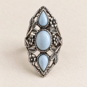May include: A silver-toned ring featuring an elongated design with three smooth, light blue oval and teardrop-shaped stones. The metal frame is decorated with detailed floral and leaf motifs, showcasing a textured, oxidized finish throughout.