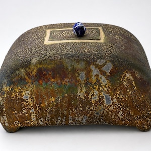 May include: A rectangular ceramic box with a textured, mottled brown and metallic finish. The lid features a central rectangular panel with an intricate pattern and a small, round, blue and white decorative bead handle centred on top.
