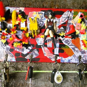 Op de afbeelding: Een rechthoekige sleutelhouder voor aan de muur met een collage van rode, zwarte en witte stukjes papier. Het bevat een miniatuurfiguurtje in anime-stijl, een gele rugzak en rode laarzen. Vier metalen haken onderaan houden diverse sleutels vast.