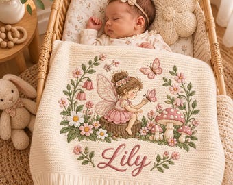 May include: A cream-colored knit baby blanket featuring an embroidered design of a fairy sitting on a tree stump surrounded by flowers, mushrooms, and butterflies. The name Lily is embroidered in pink script at the bottom of the blanket.