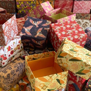 Peut inclure: Une collection de boîtes carrées en papier présentant divers motifs, notamment des imprimés floraux orange, des motifs de grues bleu foncé, des fleurs de cerisier rouges et des dessins d'éléphants beige. Les boîtes sont ouvertes et fermées.