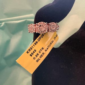 May include: A silver ring featuring three circular clusters of small diamonds. A yellow tag is attached to the band with the text KR31196-DP28, $543, 0.25 CTS, and SILVER, NATURAL DIAMOND printed in black ink on the surface of the tag.