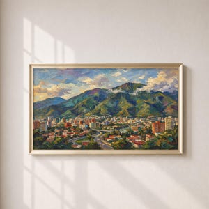 Op de afbeelding: Een olieverfschilderij in een gouden lijst met een stad aan de voet van een groot, groen gebergte onder een blauwe lucht met witte wolken. Het landschap toont talloze gebouwen met rode daken, kronkelende wegen en weelderige begroeiing.