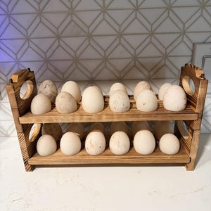 May include: A two-tiered wooden egg rack with a burnt finish, featuring circular cutouts on the side panels. The rack holds numerous off-white and speckled eggs arranged in two rows, with six eggs on the top shelf and six on the bottom shelf.