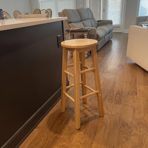 May include: A tall, light-colored wooden bar stool featuring a round seat and four straight legs connected by horizontal support rungs. The stool has a smooth, natural wood grain finish and a simple, functional design with no backrest or armrests.