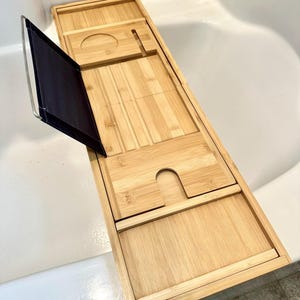 Peut inclure: Un plateau de baignoire rectangulaire en bambou doté d'un support de lecture en tissu foncé avec un cadre métallique. Le plateau comprend une découpe circulaire pour un verre, une fente rectangulaire pour un téléphone et une surface rainurée.