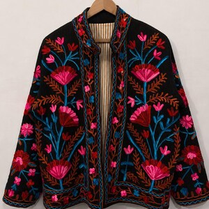 May include: A black jacket featuring vibrant floral embroidery in shades of hot pink, red, and bright blue. The garment has a mandarin collar, long sleeves, and an open front design that reveals a beige and white vertical striped lining inside.