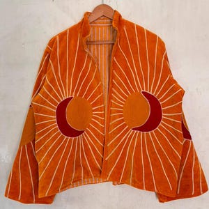 May include: An orange jacket featuring a sun and crescent moon design on each side. The suns are embroidered in a darker orange with red crescent moons, surrounded by white radiating lines. The jacket has long sleeves and an open front.