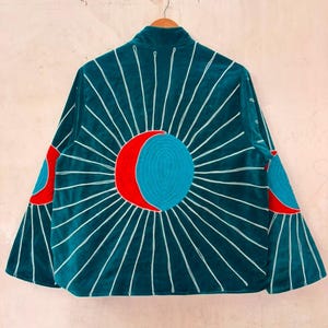 Puede incluir: Una chaqueta de terciopelo verde azulado oscuro con un gran motivo bordado de sol y luna en la espalda. El diseño incluye un círculo azul central con una media luna roja, rodeado de líneas blancas radiantes en la espalda y las mangas.