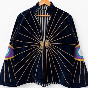 Peut inclure: Une veste en velours bleu marine foncé ornée de rayons de soleil brodés en fil doré partant du centre. Les manches présentent des motifs d'yeux circulaires brodés en violet, bleu et blanc avec des rayons dorés. La doublure est rayée noir et blanc.