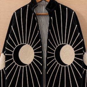 May include: A black jacket featuring a prominent cream-colored embroidered sun and moon motif on the front panels. The design includes radiating rays extending outward. The interior lining displays a vertical black and white pinstripe pattern.