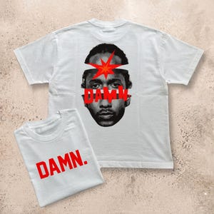 Op de afbeelding: Een lichtgrijs t-shirt met korte mouwen met een zwart-wit portret van een persoon met een rode ster en de rode tekst DAMN. over het gezicht. Een kleiner opgevouwen t-shirt op de voorgrond toont de rode tekst DAMN. op de voorzijde.