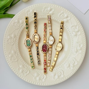 May include: Five gold-tone wristwatches with oval faces and link bracelets. The watches feature various dial colors including green, red, and white, with decorative patterned links on the bracelets displaying symbols like hearts, stars, and playing cards.