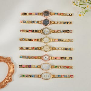 May include: Eight gold-tone link watches arranged vertically. Each watch features a unique charm-style band with colorful enamel squares depicting various symbols, stars, and hearts. The watch faces vary in color, including black and white.