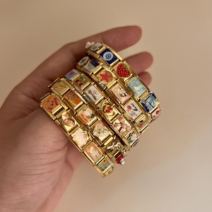 Könnte beinhalten: Ein Stapel goldfarbener Gliederarmbänder mit verschiedenen bunten Emaille-Anhängern, darunter eine Erdbeere, ein Seestern, ein böser Blick, ein Mond, Kirschen sowie Schriftzüge wie Princess, Be Mine und Bling Bling, verbunden durch Metallglieder.