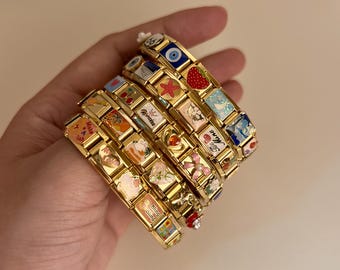 Op de afbeelding: Een stapel goudkleurige modulaire schakelarmbanden met diverse kleurrijke emaille bedels, waaronder een aardbei, zeester, boze oog, maan, kersen en teksten zoals Princess, Be Mine en Bling Bling, verbonden door in elkaar grijpende metalen schakels.