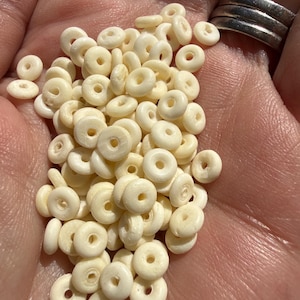 Puede incluir: Una colección de pequeñas cuentas circulares de color blanco roto con un orificio central. Cada cuenta presenta una superficie lisa y redondeada con una forma de rosquilla aplanada y consistente. Las cuentas son uniformes en color y tamaño, ofreciendo un diseño simple y funcional para diversas aplicaciones de artesanía o joyería.