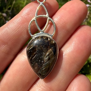 May include: A teardrop-shaped pendant featuring a dark brown and black stone with metallic inclusions, set in a polished silver-colored metal frame. The top of the pendant displays an openwork design with three interconnected curved loops.