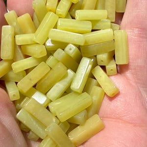 May include: A collection of small, rectangular, pale yellow beads with a smooth, polished finish. Each bead features a single, centered hole drilled through the width, creating a uniform shape suitable for stringing in various jewelry designs.
