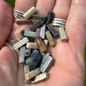 May include: A collection of small, rectangular stone beads in various shades of gray, tan, brown, and cream. Each bead features a smooth, polished finish and a single drilled hole at one end, showcasing natural mineral variations and earthy tones.