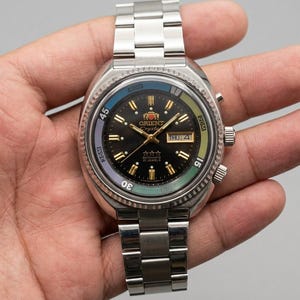 May include: A silver-tone stainless steel Orient Crystal automatic watch with a black dial, gold-tone indices, and a day-date window. The bezel features a multi-colored ring with blue, green, and light blue sections, paired with a metal link bracelet.