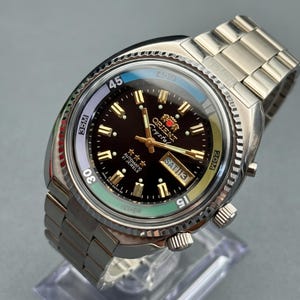 May include: An Orient Crystal automatic wristwatch with a dark brown dial, gold-tone hands and indices, and a day-date window at the three o'clock position. The watch features a silver-tone stainless steel link bracelet and a rotating bezel.