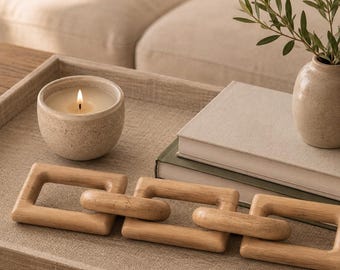 May include: A decorative wooden chain link object consisting of three interconnected square-shaped links with a smooth, light brown finish, displayed on a rectangular tray alongside a lit candle in a speckled ceramic bowl and two books.