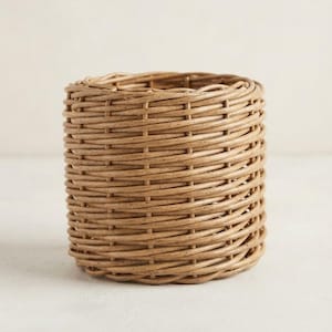 Puede incluir: Una cesta tejida cilíndrica con un acabado natural de color marrón claro y un patrón de tejido apretado y consistente. La estructura está abierta por arriba y por abajo, mostrando una forma circular uniforme hecha de material flexible.