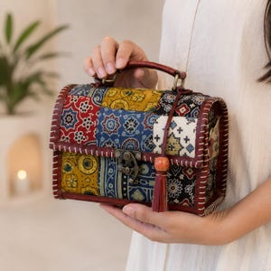 May include: A rectangular handbag featuring a patchwork design of various patterned fabrics in red, yellow, blue, and white. The bag has a dark brown leather-like handle, white stitched edges, a metal latch closure, and a small decorative tassel.