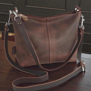 May include: A dark brown leather shoulder bag featuring a smooth matte finish, a central vertical seam, and a long adjustable strap. The bag is secured with silver-toned metal hardware, including swivel snap hooks and rivets at the strap attachment points.
