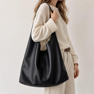 May include: A large, black hobo-style shoulder bag with a pebbled texture. The bag features a wide, integrated strap that rests comfortably on the shoulder, a spacious main compartment, and a soft, slouchy silhouette that hangs vertically.