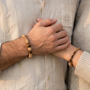 Könnte beinhalten: Zwei Perlenarmbänder an den Handgelenken. Ein Armband besteht aus flachen, hellen Holzscheibenperlen, die mit runden, braun-goldenen Tigeraugenperlen kombiniert sind. Das zweite Armband besteht aus kleineren, runden braun-goldenen Tigeraugenperlen.