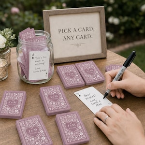 May include: A clear glass jar filled with mauve playing cards featuring a white floral heart design, alongside a framed sign reading PICK A CARD, ANY CARD. Several card decks are arranged on a table with a black marker and handwritten notes.