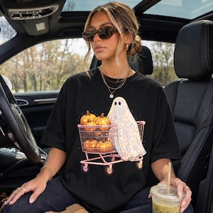 Könnte beinhalten: Ein schwarzes Kurzarm-T-Shirt mit einem Grafikdruck eines weißen Geistes mit schimmernder Textur, der einen rosa Einkaufswagen voller orangefarbener Kürbisse schiebt. Der Geist hat zwei schwarze Augen und einen kleinen Mund, und der Wagen hat vier Räder.
