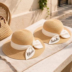 May include: Two tan straw hats with wide brims and rounded crowns. Each hat features a white fabric ribbon tied in a bow around the base of the crown. The ribbon on the left hat is embroidered with the name Olivia, and the right hat with Isabella.
