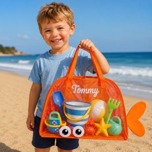 Può includere: Una borsa da spiaggia arancione in rete trasparente a forma di pesce con il nome Tommy stampato in bianco. La borsa contiene un secchiello a righe blu e bianche, una paletta blu, una palla gialla e bianca, un rastrello verde, una barchetta, un granchio e una stella marina.