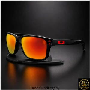 May include: Black sunglasses featuring rectangular frames and vibrant orange mirrored lenses. The temples display a prominent red Oakley logo near the hinges, with two small red circular accents on the frame front near the temple connection.
