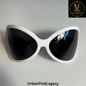 May include: Oversized sunglasses featuring a thick, smooth white plastic frame with a rounded, butterfly-inspired shape and dark, solid black lenses. The design includes a central bridge connecting the two large, curved eye pieces.