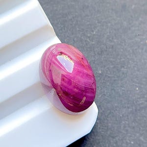 May include: An oval-shaped cabochon gemstone featuring a vibrant magenta and deep pink color palette with visible internal striations and a smooth, polished surface that reflects light, showcasing a translucent quality throughout the stone.