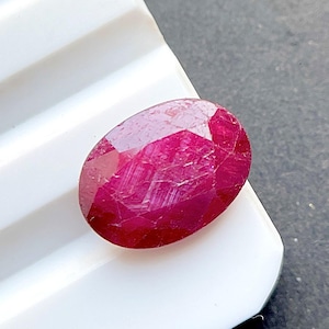 May include: An oval-shaped, deep magenta-red gemstone with a faceted surface. The stone displays a translucent quality with visible internal inclusions and a polished finish, reflecting light across its multiple cut geometric facets.