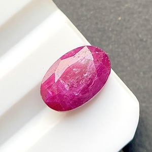 May include: An oval-shaped, faceted gemstone with a deep, vibrant magenta-red color. The stone features a polished surface with multiple geometric facets that reflect light, showcasing a translucent quality and a rich, saturated hue.