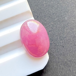 Puede incluir: Una piedra preciosa rosa de forma ovalada pulida con una superficie lisa estilo cabujón. La piedra muestra un tono rosa vibrante y translúcido con texturas cristalinas internas y un brillo reflectante en su parte superior redondeada.
